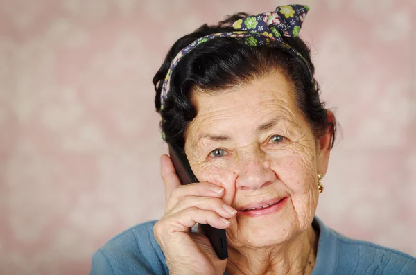 Maggiore ispanico carino donna con fiocco modello di fiore sulla testa indossando maglione blu davanti rosa carta da parati parlando in nero cellulare — Foto Stock