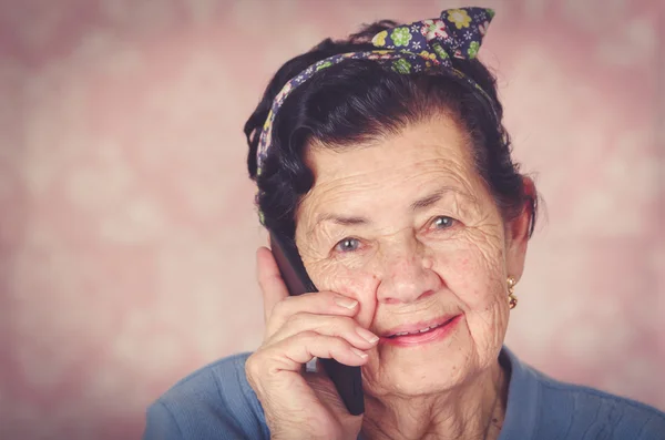 Maggiore ispanico carino donna con fiocco modello di fiore sulla testa indossando maglione blu davanti rosa carta da parati parlando in nero cellulare — Foto Stock