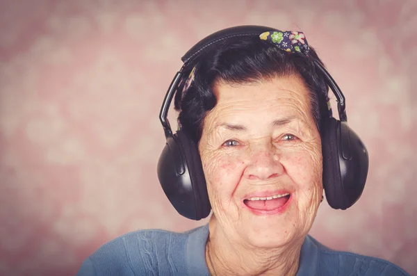 Ancienne adorable femme hispanique portant un pull bleu, motif de fleurs arc sur la tête en face de papier peint rose avec de grandes cache-oreilles noirs souriant à la caméra — Photo