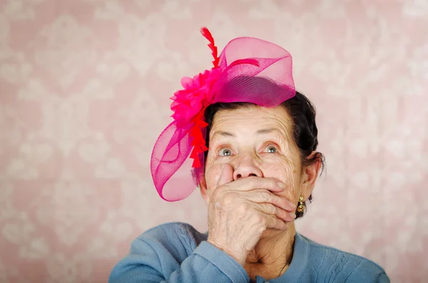 Older cute hispanic woman wearing blue sweater, large pink ribbon on head loking into camera covering her mouth with one hand