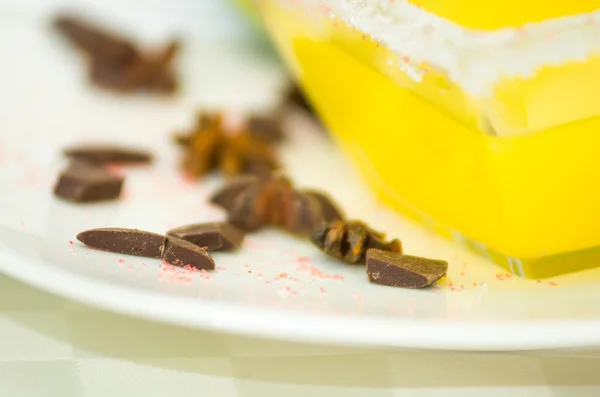 Closeup pequenos pedaços de chocolate escuro deitado na placa branca ao lado de tigela quadrada transparente açucarada com sobremesa amarela — Fotografia de Stock