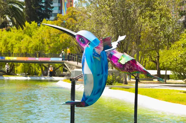 Färgglada mockingbird konstinstallation placeras på stolpar släpper vatten laguna, omgiven av träd, vacker solig dag — Stockfoto