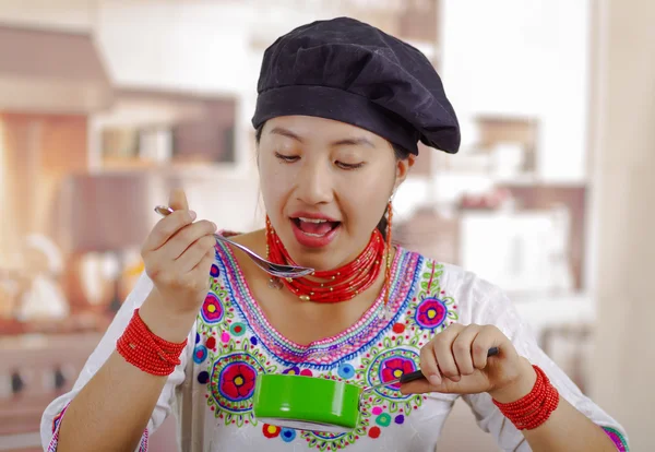 Mujer joven chef con blusa tradicional andina y sombrero de cocina, sosteniendo olla verde con salsa, degustación con cuchara, fondo de cocina —  Fotos de Stock