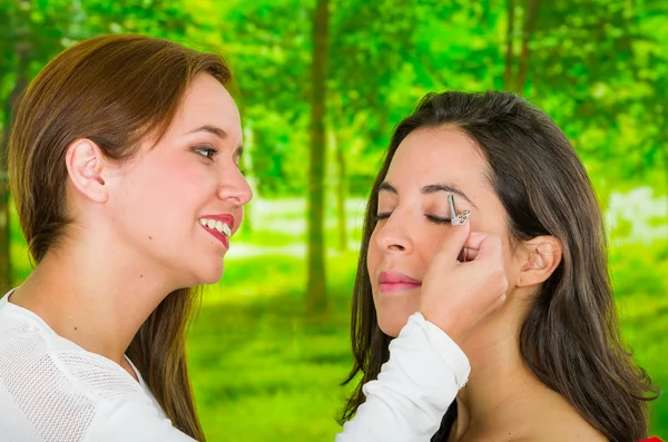 Woman performing eyebrow hair removal using tweezers on clients face, uncomfortable facial expressions, green garden background — Stock Photo, Image
