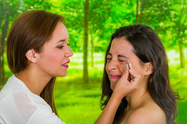 Woman performing eyebrow hair removal using tweezers on clients face, uncomfortable facial expressions, green garden background — Stock Photo, Image