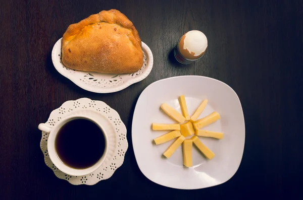 Petit déjeuner d'inspiration latine sur une table en bois sombre, y compris des tranches de fromage placées sur une assiette carrée blanche à côté du verre de jus d'orange, du café, des œufs durs et un pain empanada — Photo