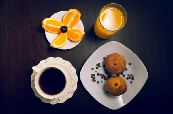 Petit déjeuner sur une surface en bois sombre comprenant café, muffins, jus d'orange et tranches d'oranges placées sur une petite assiette blanche — Photo