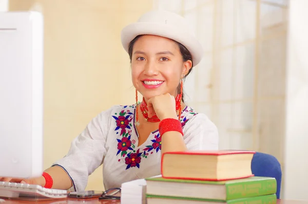 Giovane bella ragazza che indossa camicia bianca con decorazioni floreali colorate e cappello alla moda sorridente, seduto alla scrivania, pila di libri, sfondo luminoso — Foto Stock