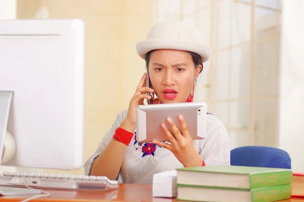 Giovane bella ragazza indossa camicia bianca con decorazioni floreali colorate e cappello alla moda, seduto alla scrivania guardando schermo tablet parlare al telefono, pila di libri, sfondo luminoso — Foto Stock