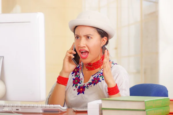 Joven chica bonita con camisa blanca con decoraciones de flores de colores y sombrero de moda, sentado junto al escritorio hablando por teléfono expresión facial molesta, pila de libros, fondo brillante — Foto de Stock