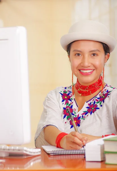 Young pretty girl wearing white shirt with colorful flower decorations and fashionable hat, sitting by desk working writing on paper smiling, stack of books, bright background — Stock Photo, Image