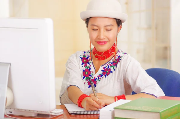Giovane bella ragazza che indossa camicia bianca con decorazioni floreali colorate e cappello alla moda, seduta alla scrivania a lavorare scrivendo su carta sorridente, pila di libri, sfondo luminoso — Foto Stock