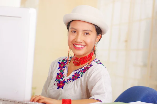 Giovane bella ragazza che indossa camicia bianca con decorazioni floreali colorate e cappello alla moda, seduta alla scrivania a lavorare scrivendo su carta sorridente, pila di libri, sfondo luminoso — Foto Stock