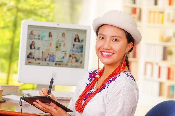 Giovane bella ragazza con camicia bianca e cappello alla moda, seduta alla scrivania del computer, girando verso la fotocamera sorridente felicemente — Foto Stock