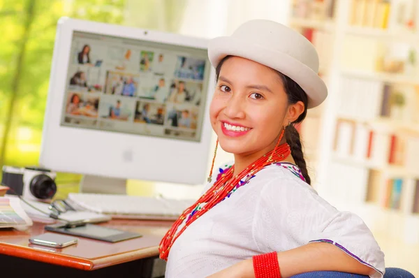 Giovane bella ragazza con camicia bianca e cappello alla moda, seduta alla scrivania del computer, girando verso la fotocamera sorridente felicemente — Foto Stock