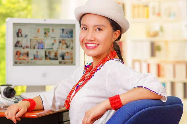 Jong mooi meisje dragen wit shirt en modieuze hoed, zitten door computer bureau, draaien in de richting van camera glimlachen gelukkig — Stockfoto