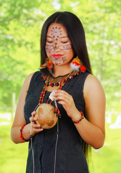 Femme amazonienne avec peinture faciale et robe noire posant pour appareil photo, tenant une bouteille en bois, fond de forêt — Photo