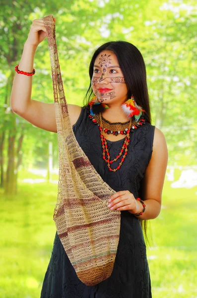 Mulher exótica amazônica com pintura facial e vestido preto, saco natural pendurado na parte superior do corpo, posando para câmera, fundo da floresta — Fotografia de Stock