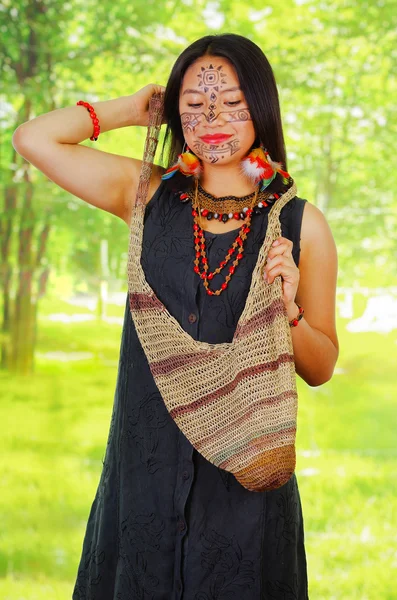 Mujer exótica amazónica con pintura facial y vestido negro, bolsa natural colgando en la parte superior del cuerpo, posando para la cámara, fondo del bosque —  Fotos de Stock