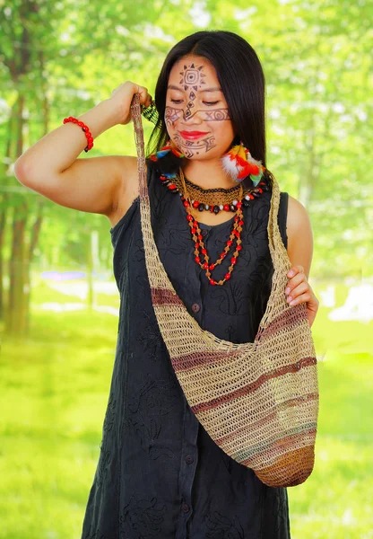Femme exotique amazonienne avec peinture faciale et robe noire, sac naturel suspendu sur le haut du corps, posant pour appareil photo, fond de forêt — Photo