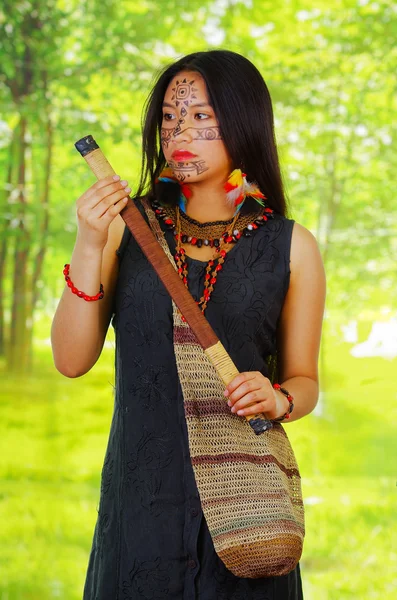 Femme exotique amazonienne avec peinture faciale et robe noire, sac naturel suspendu sur le haut du corps, tirant sur la sarbacane, posant pour appareil photo, fond de forêt — Photo