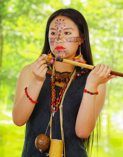 Εξωτική γυναίκα του Αμαζονίου, βαφή προσώπου, μαύρο φόρεμα, θήκη για βέλη και μικρό ξύλινο μπουκάλι που κρέμεται γύρω από το λαιμό, φορτώνοντας το βέλος στο φυσικοπίστολο, δάσος φόντο. — Φωτογραφία Αρχείου
