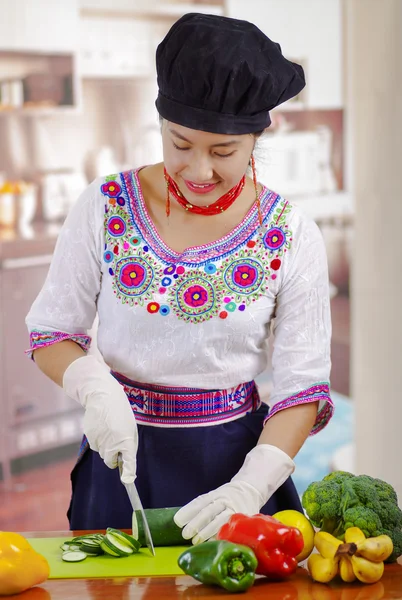 Giovane donna chef indossa camicetta andina tradizionale e cappello da cucina, verdure sulla scrivania, usando coltello tagliare fette di cetriolo, fondo cucina — Foto Stock