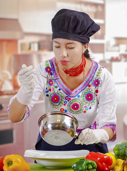 Giovane chef indossa camicetta andina tradizionale e cappello da cucina, verdure sulla scrivania, cucchiaio in mano, pentola da cucina in altra mano, sfondo cucina — Foto Stock