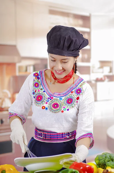 Giovane chef indossa camicetta andina tradizionale, cappello da cucina nero, verdure sulla scrivania, tenendo piatto profondo e frusta in altra mano, sfondo della cucina — Foto Stock