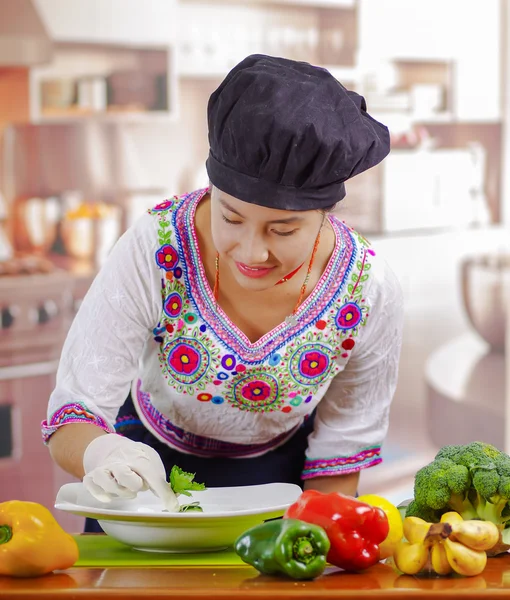 Young woman chef wearing traditional andean blouse, black cooking hat, vegetables on desk, placing green leaf inside deep plate, kitchen background — Stock Photo, Image