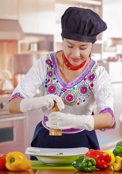 Giovane donna chef indossa camicetta andina tradizionale, cappello da cucina nero, verdure sulla scrivania, utilizzando macinapepe su piatto profondo, fondo cucina — Foto Stock