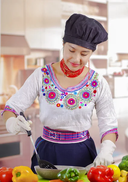Jovem chef vestindo blusa tradicional andina, chapéu de cozinha preto, legumes na mesa, usando frigideira e espátula, fundo da cozinha — Fotografia de Stock