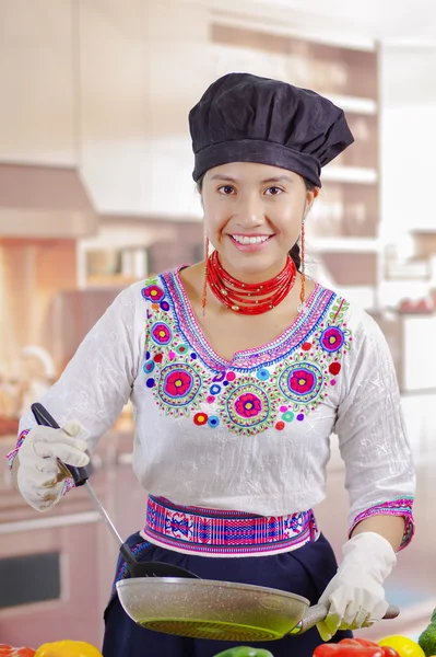 Jonge vrouw chef-kok met traditionele Andes blouse, zwarte kokende hoed, groenten op het bureau, met behulp van koekenpan en spatel, keuken achtergrond — Stockfoto