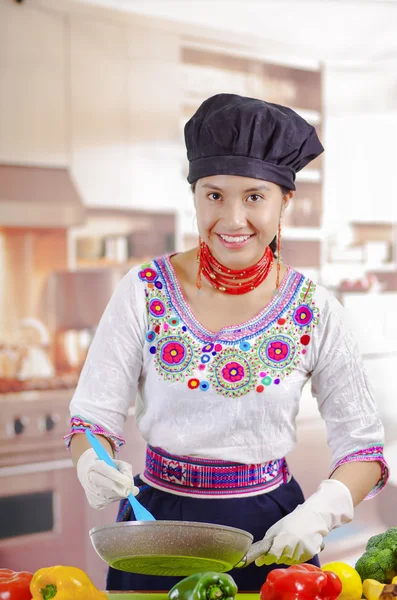 Jovem chef vestindo blusa tradicional andina, chapéu de cozinha preto, legumes na mesa, usando frigideira e espátula, fundo da cozinha — Fotografia de Stock