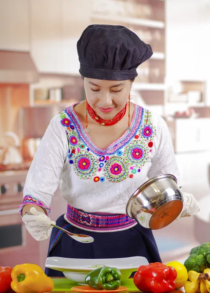 Jovem chef vestindo blusa andina tradicional e chapéu de cozinha, legumes na mesa, segurando panela de cozinhar com molho, fundo da cozinha — Fotografia de Stock