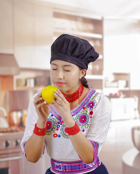 Mujer joven chef con blusa andina tradicional, sombrero de cocina negro, sosteniendo limón para la sonrisa de la cámara, fondo de cocina —  Fotos de Stock