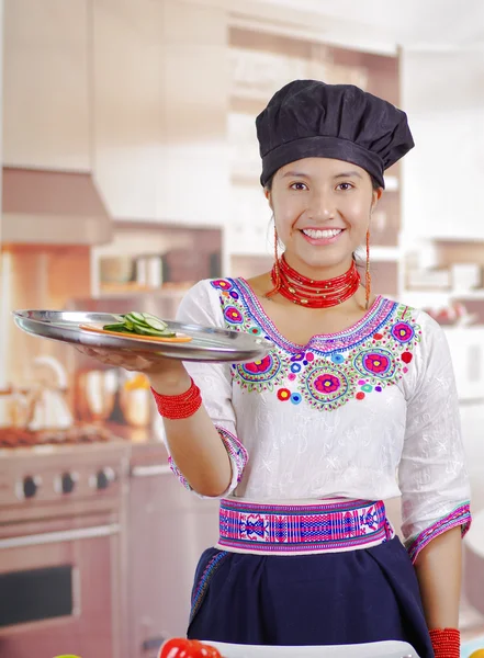 Young woman chef wearing traditional andean blouse, black cooking hat, holding up large tray with cucumber slices on top, kitchen background