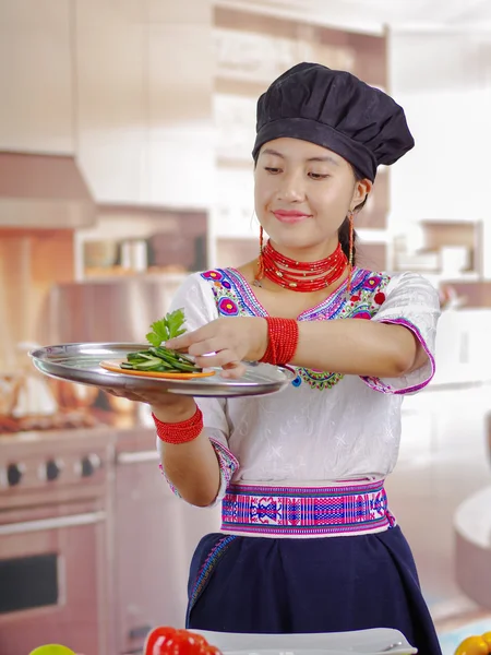 Giovane chef indossa camicetta andina tradizionale, cappello da cucina nero, tenendo in mano grande vassoio con fette di cetriolo sulla parte superiore, sfondo della cucina — Foto Stock