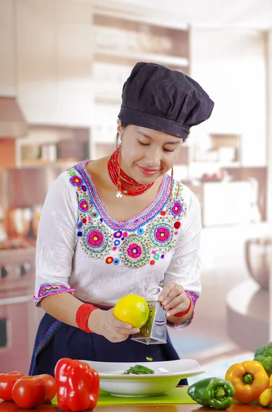 Giovane chef indossa camicetta andina tradizionale, cappello da cucina nero, verdure sulla scrivania, triturazione limone in piatto profondo, sfondo cucina — Foto Stock