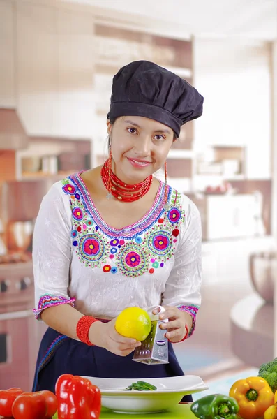 Jovem chef vestindo blusa tradicional andina, chapéu de cozinha preto, legumes na mesa, trituração de limão em placa profunda, fundo da cozinha — Fotografia de Stock