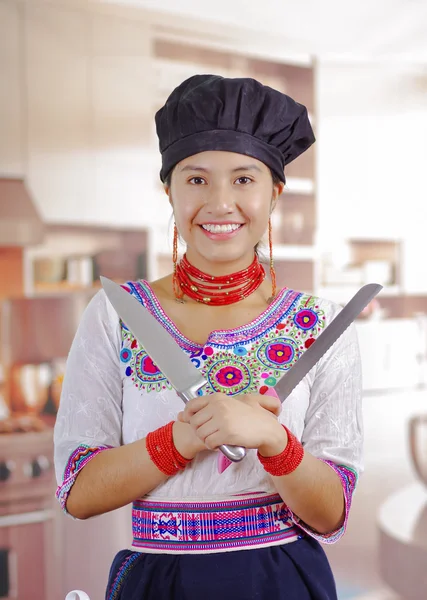 Joven chef con blusa andina tradicional, sombrero de cocina negro, sosteniendo dos cuchillos grandes, cruzándolos frente al pecho, fondo de cocina —  Fotos de Stock