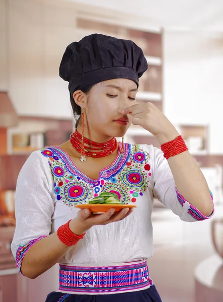 Jonge vrouw chef-kok met traditionele Andes blouse, zwarte kokende hoed, holding plaat van komkommer plakjes, bedekking neus voor slechte geur, sceptische gezichtsuitdrukking, keuken achtergrond — Stockfoto