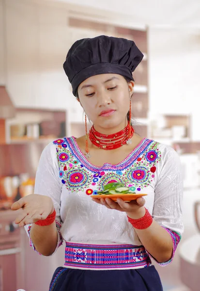Giovane donna chef indossa camicetta andina tradizionale, cappello da cucina nero, tenendo piatto di fette di cetriolo, espressione facciale scettica, fondo cucina — Foto Stock