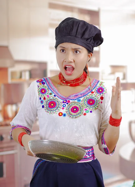 Jonge vrouw chef-kok met traditionele Andes blouse, zwarte kokende hoed, met koekenpan en kijken naar binnen met overstuur gezichtsuitdrukking, keuken achtergrond — Stockfoto