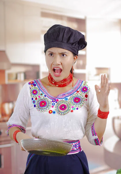 Mujer joven chef con blusa andina tradicional, sombrero de cocina negro, sosteniendo sartén y mirando dentro con expresión facial alterada, fondo de cocina —  Fotos de Stock
