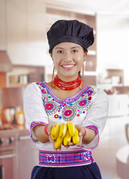 Mujer joven chef con blusa andina tradicional, sombrero de cocina negro, sosteniendo plátanos mostrando a la cámara y sonriendo, fondo de cocina — Foto de Stock