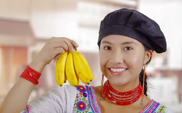 Mujer joven chef con blusa andina tradicional, sombrero de cocina negro, sosteniendo plátanos mostrando a la cámara y sonriendo, fondo de cocina —  Fotos de Stock