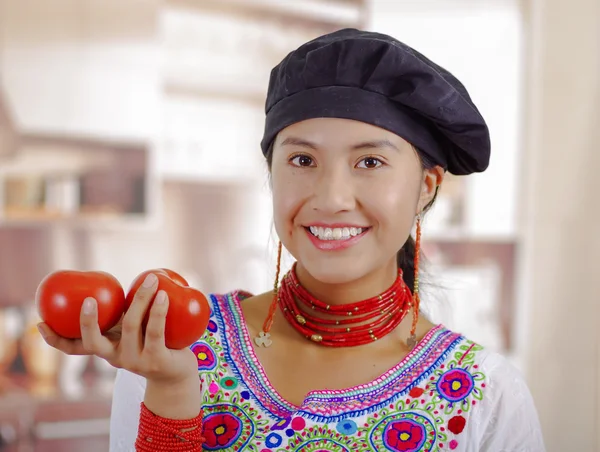 Mujer joven chef con blusa andina tradicional, sombrero de cocina negro, sosteniendo tomates mostrando a la cámara y sonriendo, fondo de cocina —  Fotos de Stock