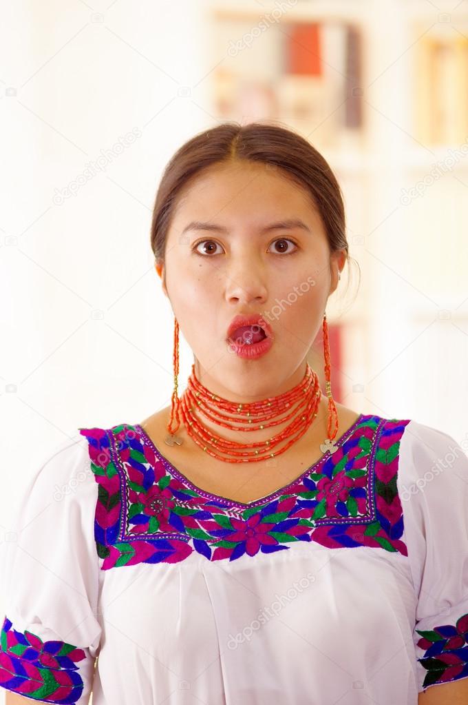 Beautiful young brunette woman wearing traditional andean white blouse with blue decorative edges, facing camera interacting surprised, bright household background