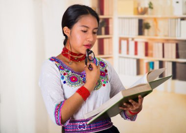 Geleneksel Andean bluzu giyen, elinde gözlük ve kitap okuyan güzel genç bir avukat, arka planda kitaplıklar.